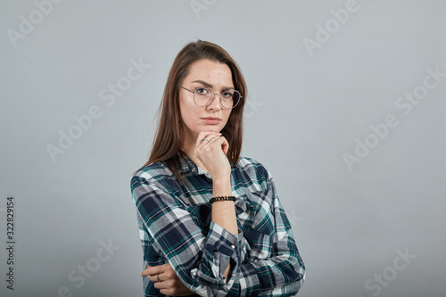 Young brunette girl blue green in checked shirt on grey background smart woman with glasses thinks, makes decisions stole her head on hand