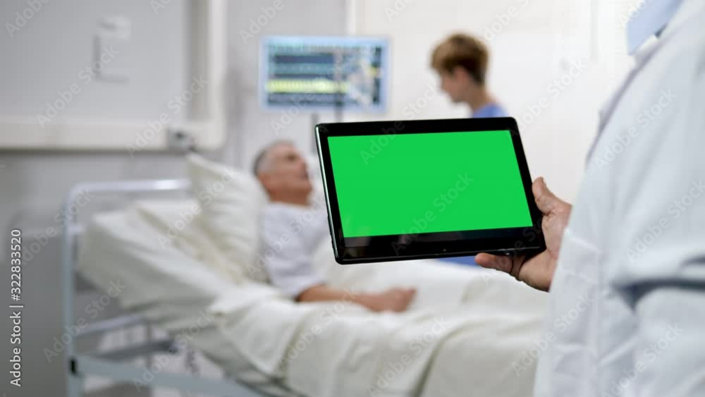 doctor holding a green screen tablet with a nurse taking care of a senior patient male lying in the hospital bed blurred in the background doctor holding a green screen tablet with a nurse taking care of a senior patient male lying in the hospital bed blurred in the background