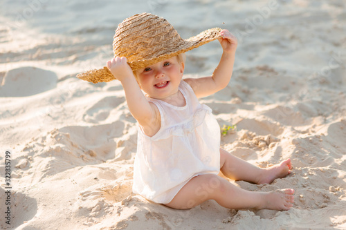 baby girl in white clothes and a straw hat sits on the white sand on the beach in summer