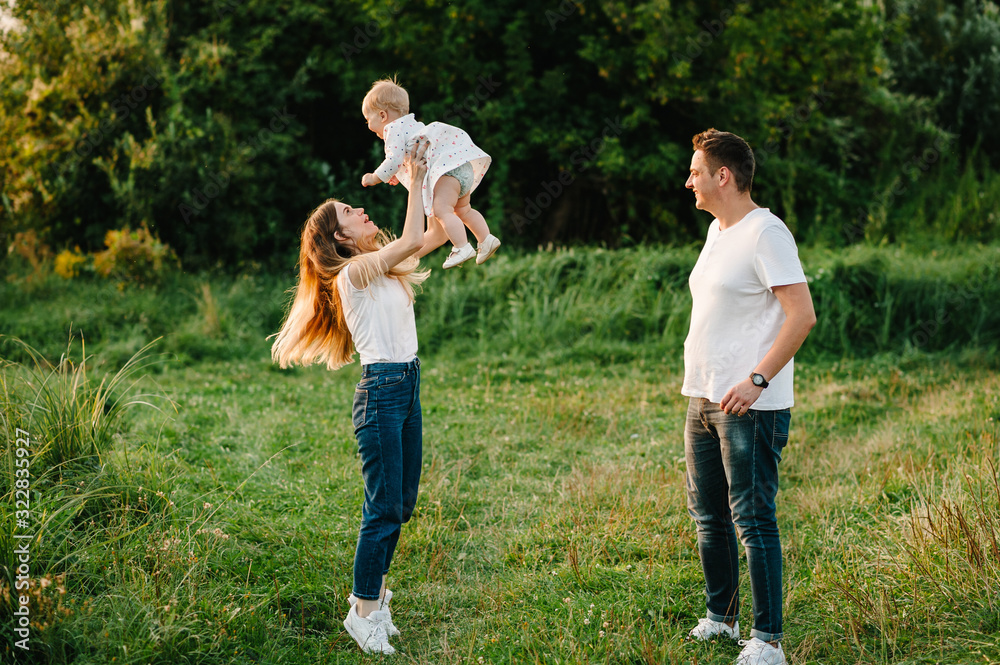 Fototapeta premium Portrait of a father, mother holds, throws up and spins the daughter on hands walking on nature on summer day vacation. Mom, dad and girl playing in the park. Concept of happy family.