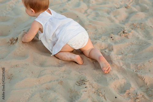 baby girl in white clothes and a straw hat sits on the white sand on the beach in summer