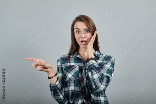Young brunette girl blue green in checked shirt on grey background shocked woman in glasses points the direction