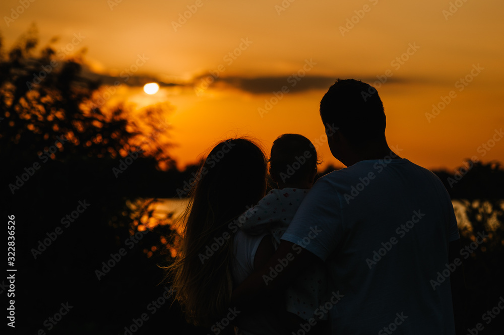 Mom, dad holds daughter stand back and looking on sunset. Portrait of a young family on nature, on vacation, outdoors.  The concept of family holiday.