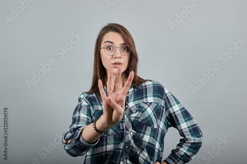 Young brunette girl blue green in checked shirt on grey background stylish woman in glasses surprised, shows four fingers