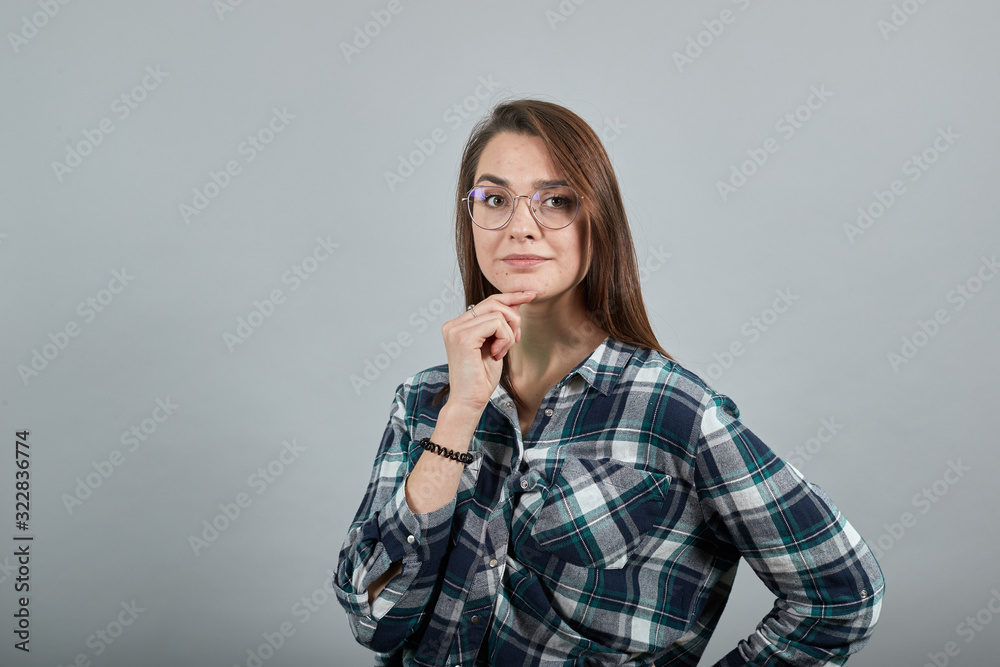 Young brunette girl blue green in checked shirt on grey background smart woman with glasses thinks, holds her chin with hand Young brunette girl blue green in checked shirt on grey background smart woman with glasses thinks, holds her chin with hand