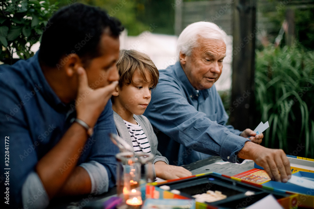 Multi-generation family playing board game while sitting at table in backyard Multi-generation family playing board game while sitting at table in backyard