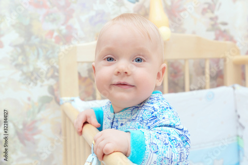 A little boy, a child with blue eyes is standing in a crib and smiling at home. Learns to stand at the support. Portrait, close-up