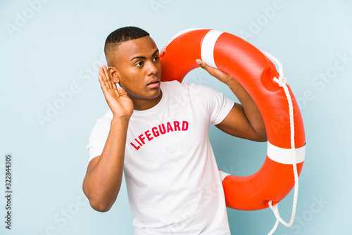 Young latin life guard isolated on blue background trying to listening a gossip. © Asier