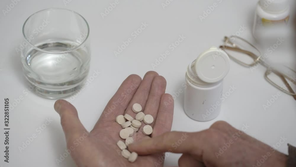 senior man with wrinkled worn-out hands takes pile of white pills from jar at table in room extreme close backside view senior man with wrinkled worn-out hands takes pile of white pills from jar at table in room extreme close backside view