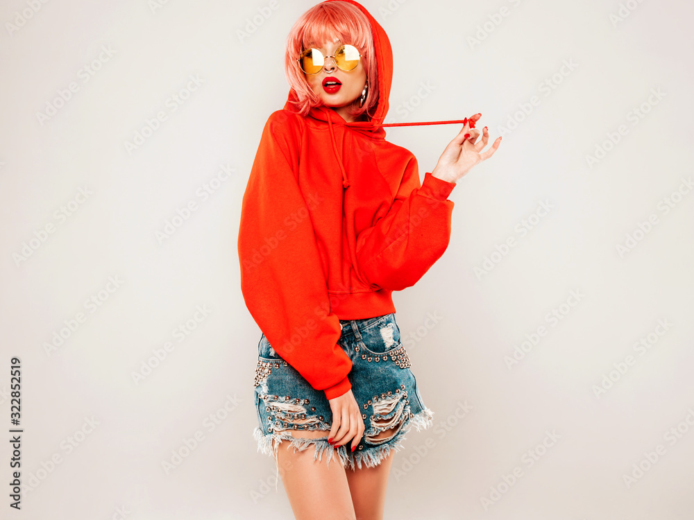 Young beautiful hipster bad girl in trendy red summer hoodie and earring in her nose.Sexy carefree woman in pink wig posing in studio on gray background.Hot model in sunglasses Young beautiful hipster bad girl in trendy red summer hoodie and earring in her nose.Sexy carefree woman in pink wig posing in studio on gray background.Hot model in sunglasses