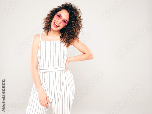 Portrait of beautiful smiling model with afro curls hairstyle dressed in summer hipster clothes.Sexy carefree girl posing in studio on gray background.Trendy funny and positive woman in sunglasses