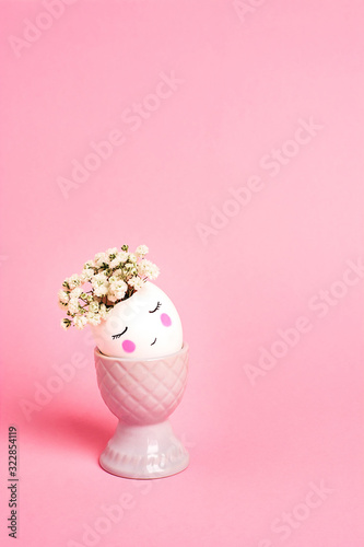 Easter creative concept. Easter decorative egg with painted cheerful faces and flowers instead of hair in the egg cup on the pink background. Copy space, close up