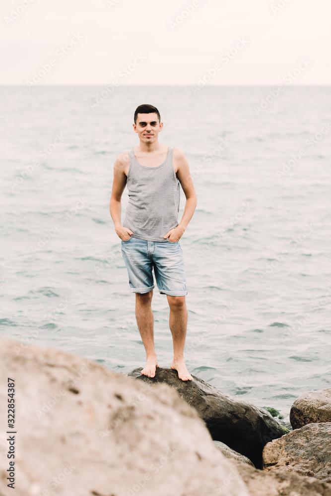 a young guy in a t-shirt and shorts stands on the beach on large rocks