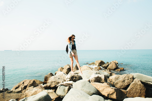 a young girl traveler with a backpack walks barefoot on large stones on the sea coast