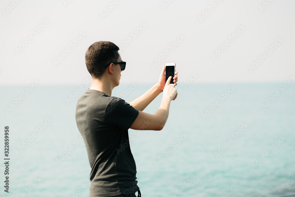 a young guy in a t-shirt and shorts on large rocks, taking pictures of a picturesque place on the sea coast on a smartphone