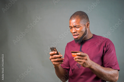 handsome young african man isolated over grey background making transaction online with his mobile phone and credit card. © Vic Josh