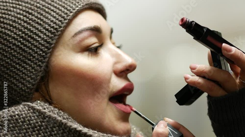 Girl in winter jacket applies makeup and paints her lips in lobby of metro station in Moscow, Russia. Young beautiful woman with lipstick brush is standing in subway and looking in pocket mirror.
