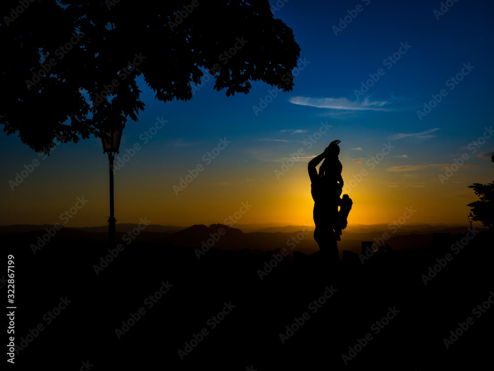 Silhouette of a statue during sunset with colorful clouds and blue sky and sun behind the sculpture