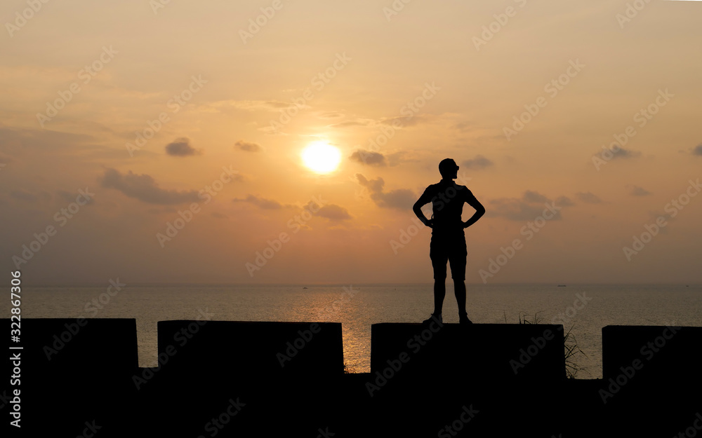 Silhouette of tall man standing on low wall against backdrop of beautiful sunset. Young traveler figure on sea background. Silhouette of tall man standing on low wall against backdrop of beautiful sunset. Young traveler figure on sea background.