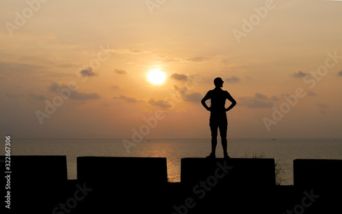 Silhouette of tall man standing on low wall against backdrop of beautiful sunset. Young traveler figure on sea background.