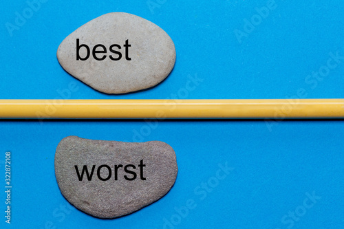 The words best and worst are written on natural smooth stones separated by a yellow pencil. The background is isolated in blue and has a lot of space photo
