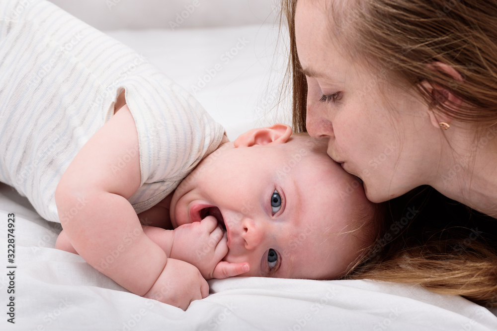 Young mother kissing baby lying on bed, close up. Lovely time together. Happy motherhood concept Young mother kissing baby lying on bed, close up. Lovely time together. Happy motherhood concept
