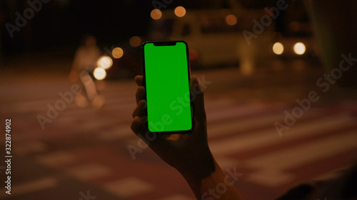 Lviv, Ukraine - May 19, 2018: Close-up of young urban girl typing message to friend online on vertical smartphone with mock-up greenscreen outdoor in the street. Night life and technology.
