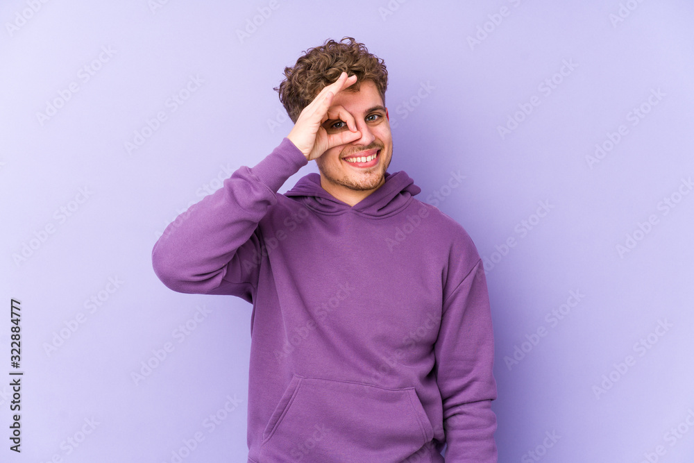 Young blond curly hair caucasian man isolated excited keeping ok gesture on eye. Young blond curly hair caucasian man isolated excited keeping ok gesture on eye.