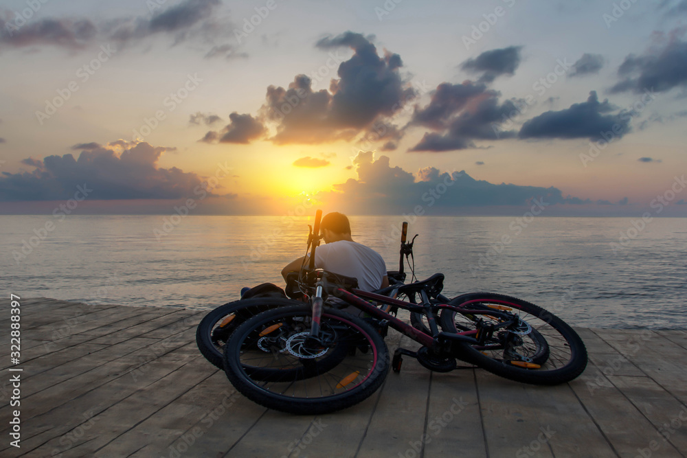 After cycling man in white short sits near sea and works or dreams or writes. View from the back. His bicycles near him. Sunset or sunrise.