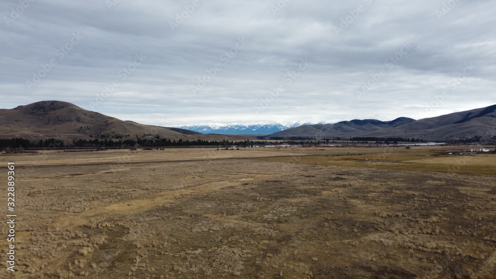 Vast open field leading to a wide river and rolling hill type mountains with a snow covered mountain range in the distance
