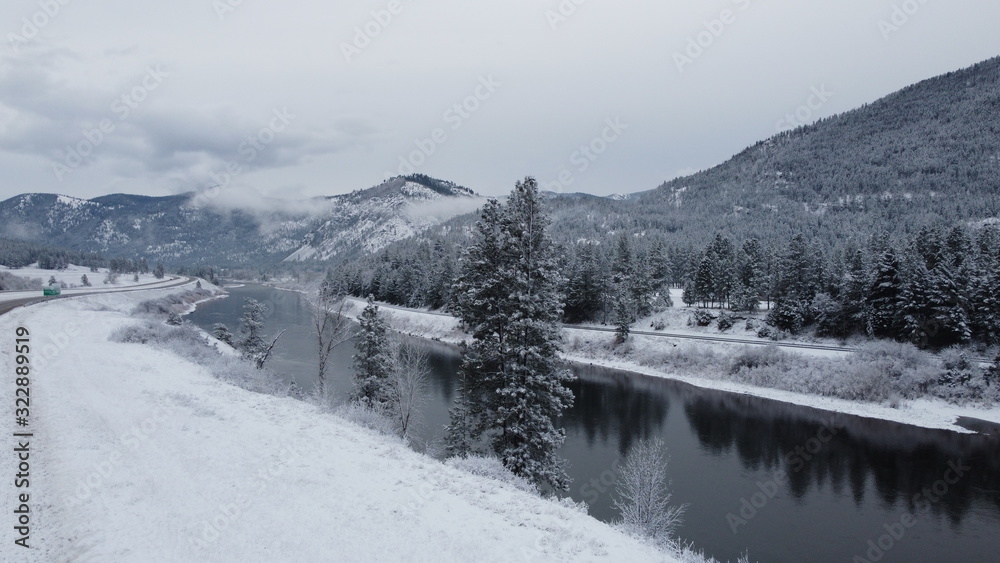 Fototapeta premium Large river cutting through snow covered valley with tree mountain range and curving roads
