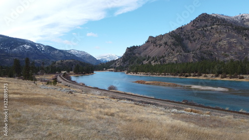 Wide river and train track cutting through large grass covered valley between tree covered rolling hills and snow covered mountains