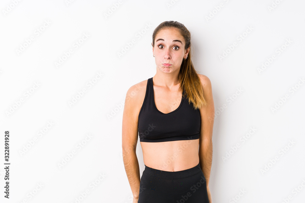 Young caucasian fitness woman posing in a white background shrugs shoulders and open eyes confused. Young caucasian fitness woman posing in a white background shrugs shoulders and open eyes confused.