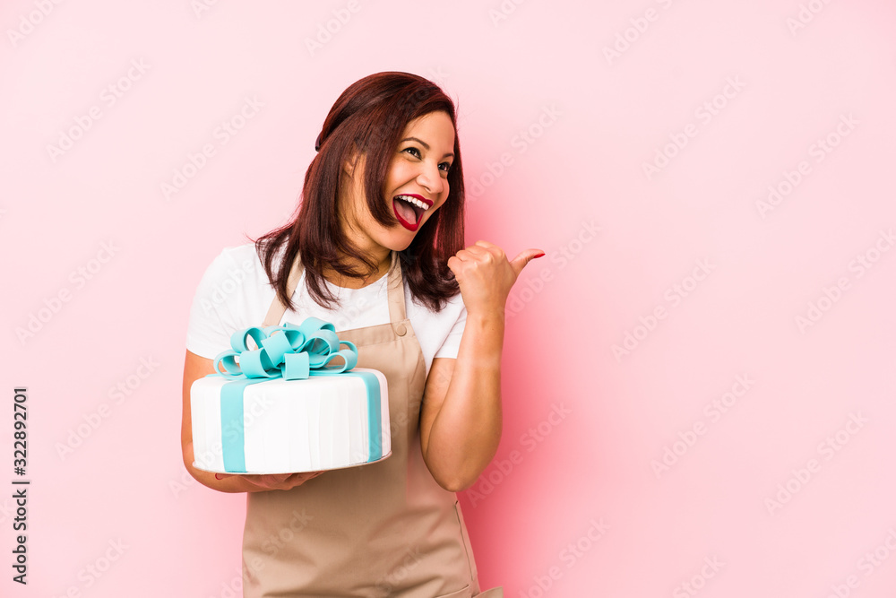 Middle age latin woman holding a cake isolated on a pink background points with thumb finger away, laughing and carefree. Middle age latin woman holding a cake isolated on a pink background points with thumb finger away, laughing and carefree.