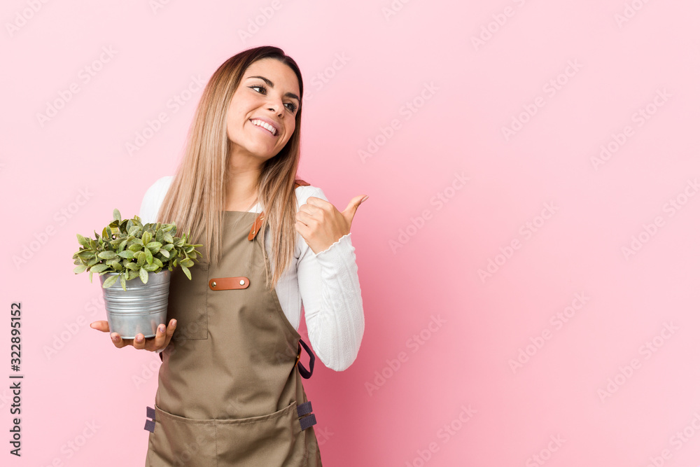 Young gardener woman holding a plant points with thumb finger away, laughing and carefree.