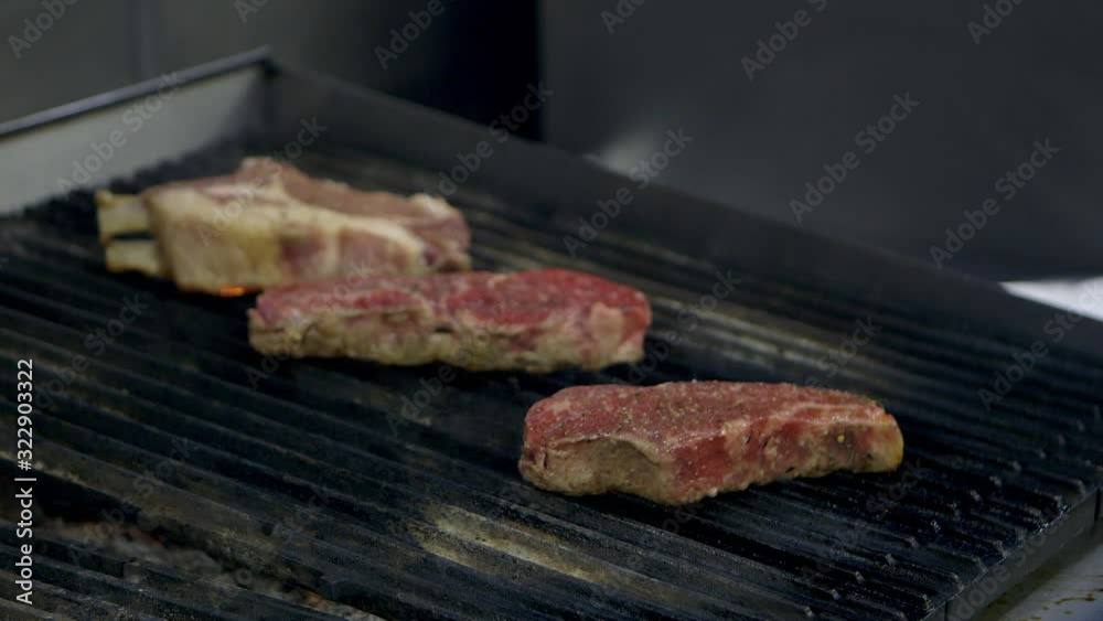 Medium close up of strip steaks and a pork chop cooking on a grill in a restaurant kitchen.