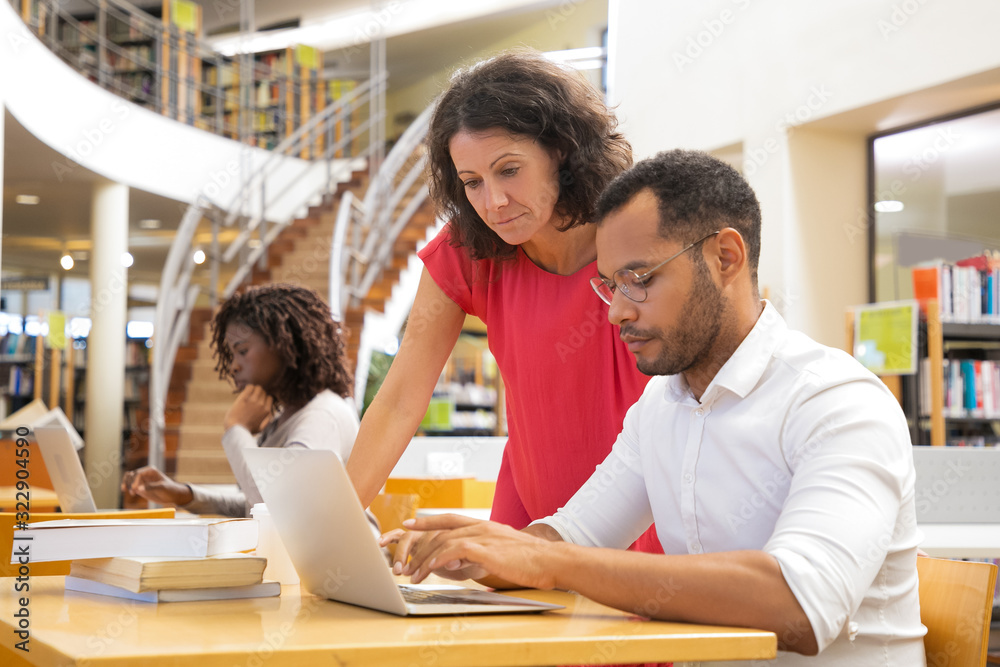 Serious colleagues working with laptop at library. Focused mature woman standing near concentrated African American man who typing on laptop. Education concept