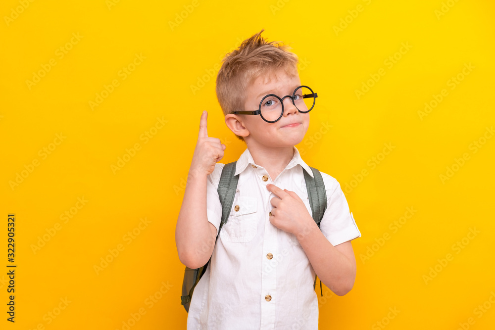 Back to school. Portrait of funny clever blonde school boy in round glasses with bag in white shirt. Pointing up. Yellow studio background. Education. Looking and smiling at camera - obrazy, fototapety, plakaty 
