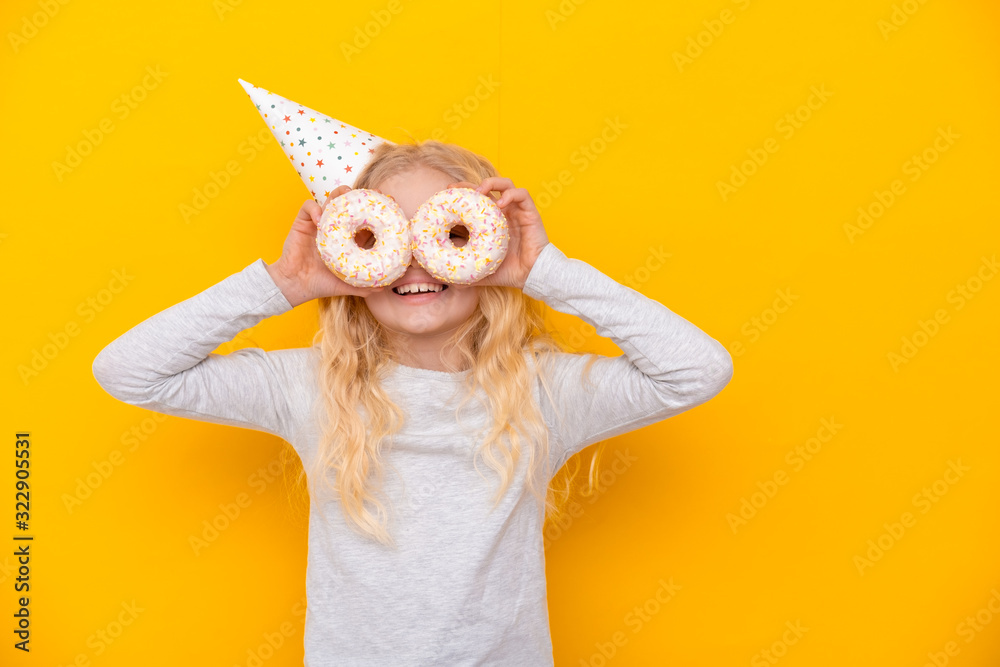 Crazy cheerful blonde girl in birthday hat smiling, having fun and looking through two white donuts on her eyes. Sweets. Yellow studio background.