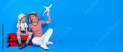 Full length happy excited woman and little boy in summer clothes and hats with plane sitting on red suitcase. Travel and tourism. isolated over blue background. Banner. Copyspace.
