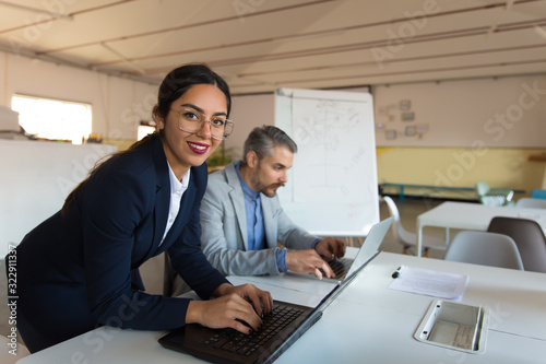 Smiling Asian businesswoman looking at camera. Cheerful young woman wearing suit typing on laptop and looking at camera. Business concept