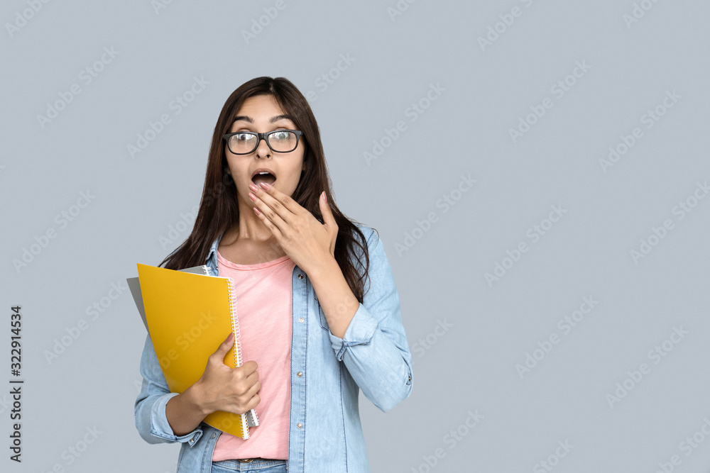 Shocked young adult indian student teen girl looking at camera, holding copybooks in hand and covering mouth with palm. Woman teenager standing isolated on grey background with copy space Shocked young adult indian student teen girl looking at camera, holding copybooks in hand and covering mouth with palm. Woman teenager standing isolated on grey background with copy space
