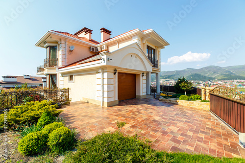 The facade of a classic Mediterranean two-storey cottage in the spring. In front of the cottage there is a green lawn and flowering trees. Mountains in the background.