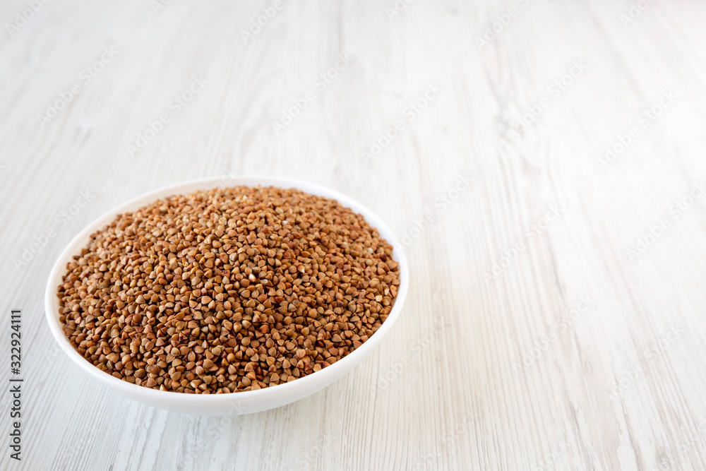 Uncooked Roasted Buckwheat in a white bowl on a white wooden surface, low angle view. Copy space. Uncooked Roasted Buckwheat in a white bowl on a white wooden surface, low angle view. Copy space.
