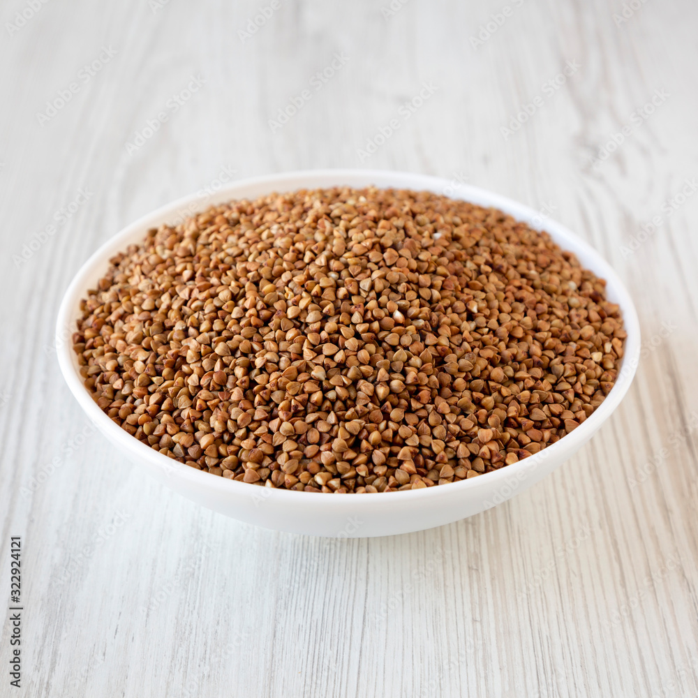 Uncooked Roasted Buckwheat in a white bowl on a white wooden table, side view. Uncooked Roasted Buckwheat in a white bowl on a white wooden table, side view.