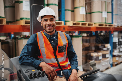 Handsome engineer wearing protective uniform and hardhat smiling joyfully to the camera sitting in forklift stacker while working at the storage