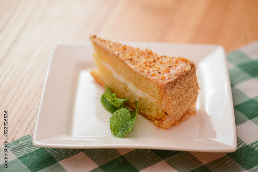 Slice of victoria sponge cake with cream filling, served with a mint leaf on a white plate on light wooden table Slice of victoria sponge cake with cream filling, served with a mint leaf on a white plate on light wooden table