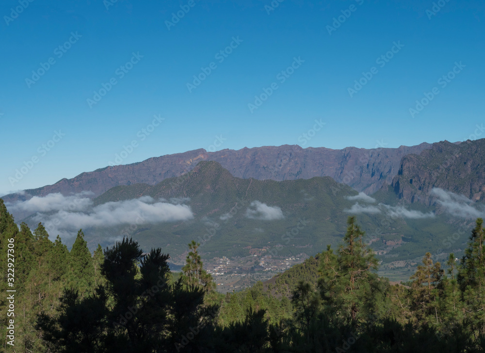Volcanic landscape with lush green pine trees, colorful volcanoes and lava rock field along path Ruta de los Volcanes, hiking trail at La Palma island, Canary Islands, Spain, Blue sky background