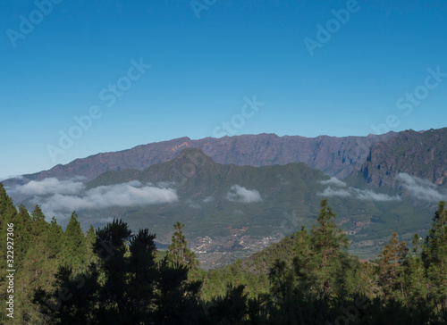 Volcanic landscape with lush green pine trees, colorful volcanoes and lava rock field along path Ruta de los Volcanes, hiking trail at La Palma island, Canary Islands, Spain, Blue sky background © Kristyna