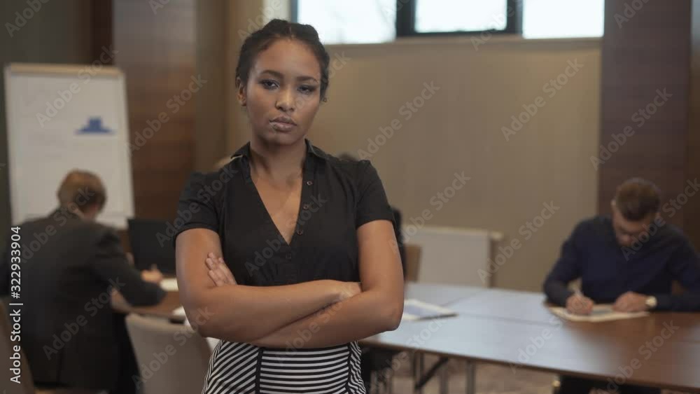 international business meeting, young african businesswoman stands in conference room and looks at the camera, serious look, colleagues works in the blurred background. international business meeting, young african businesswoman stands in conference room and looks at the camera, serious look, colleagues works in the blurred background.